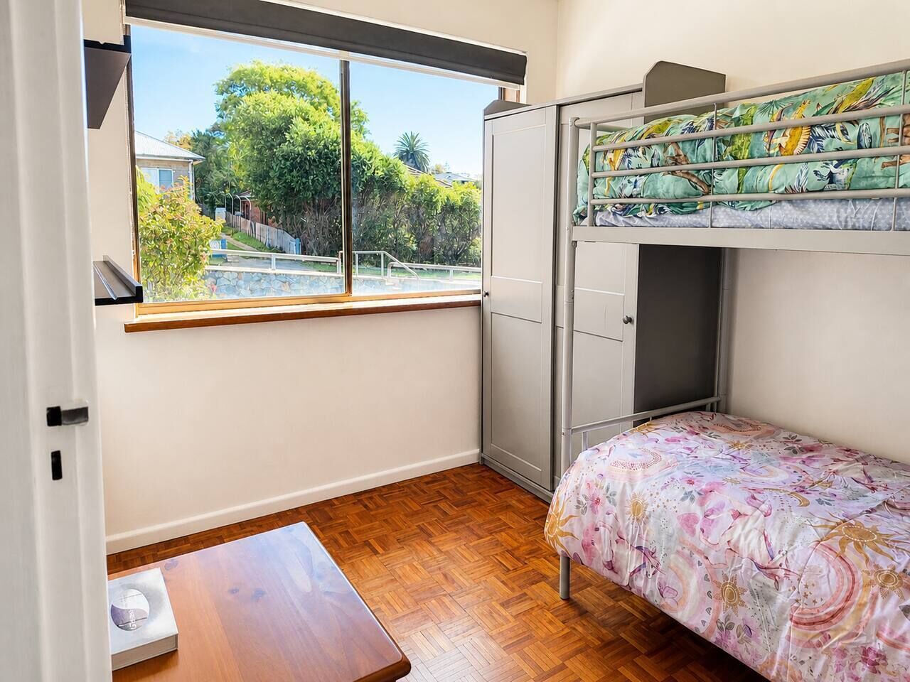Une chambre avec un lit superposé, une petite table, une fenêtre donnant sur un espace verdoyant et une porte.