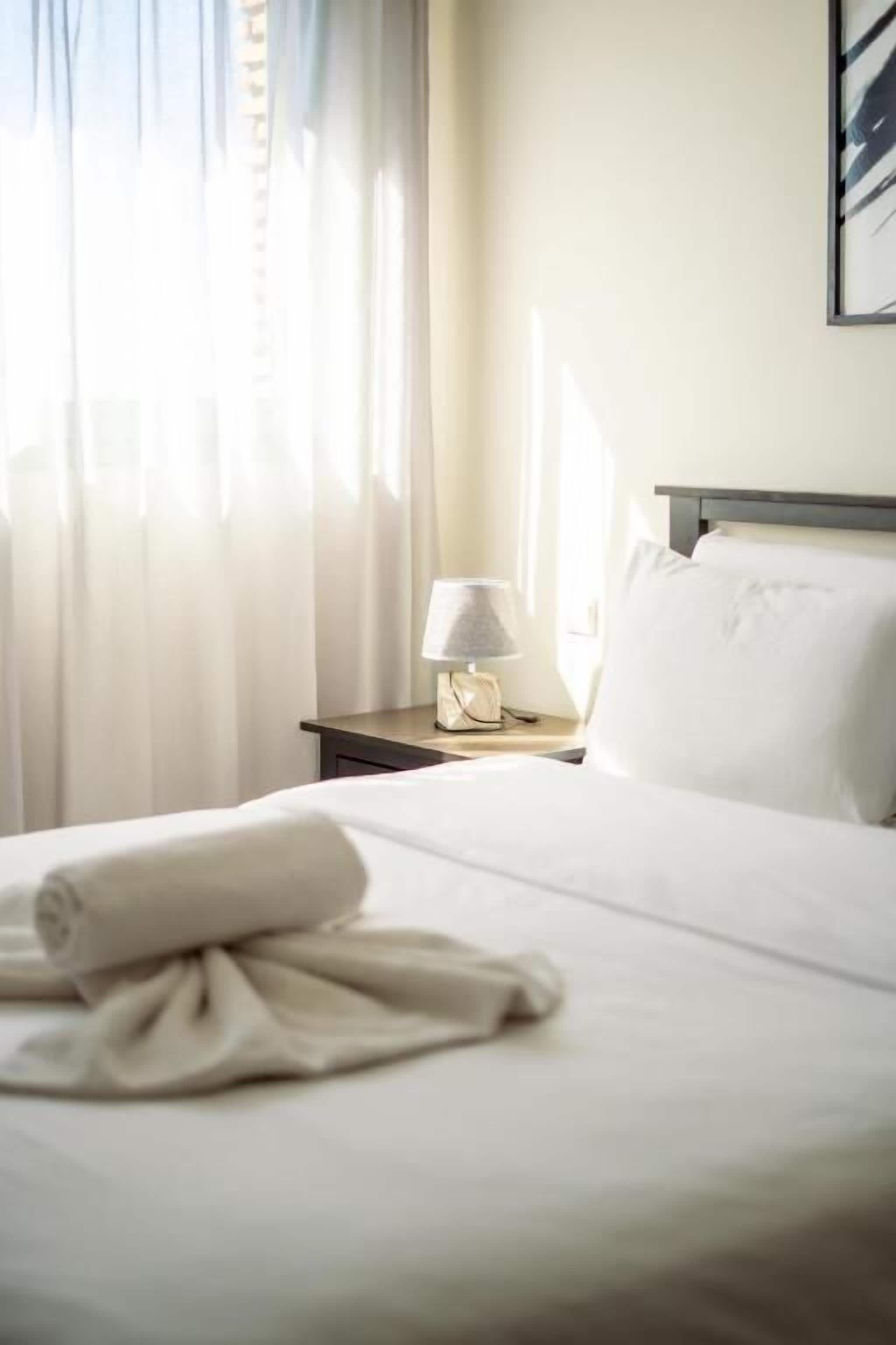 A hotel room with a neatly made bed, a bedside lamp, and a window with sheer curtains.