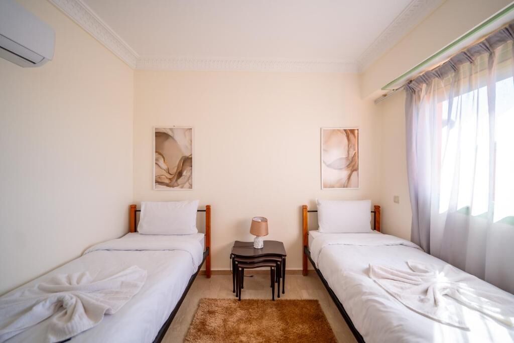 A hotel room with two single beds, a small table with a lamp, and artwork on the walls.