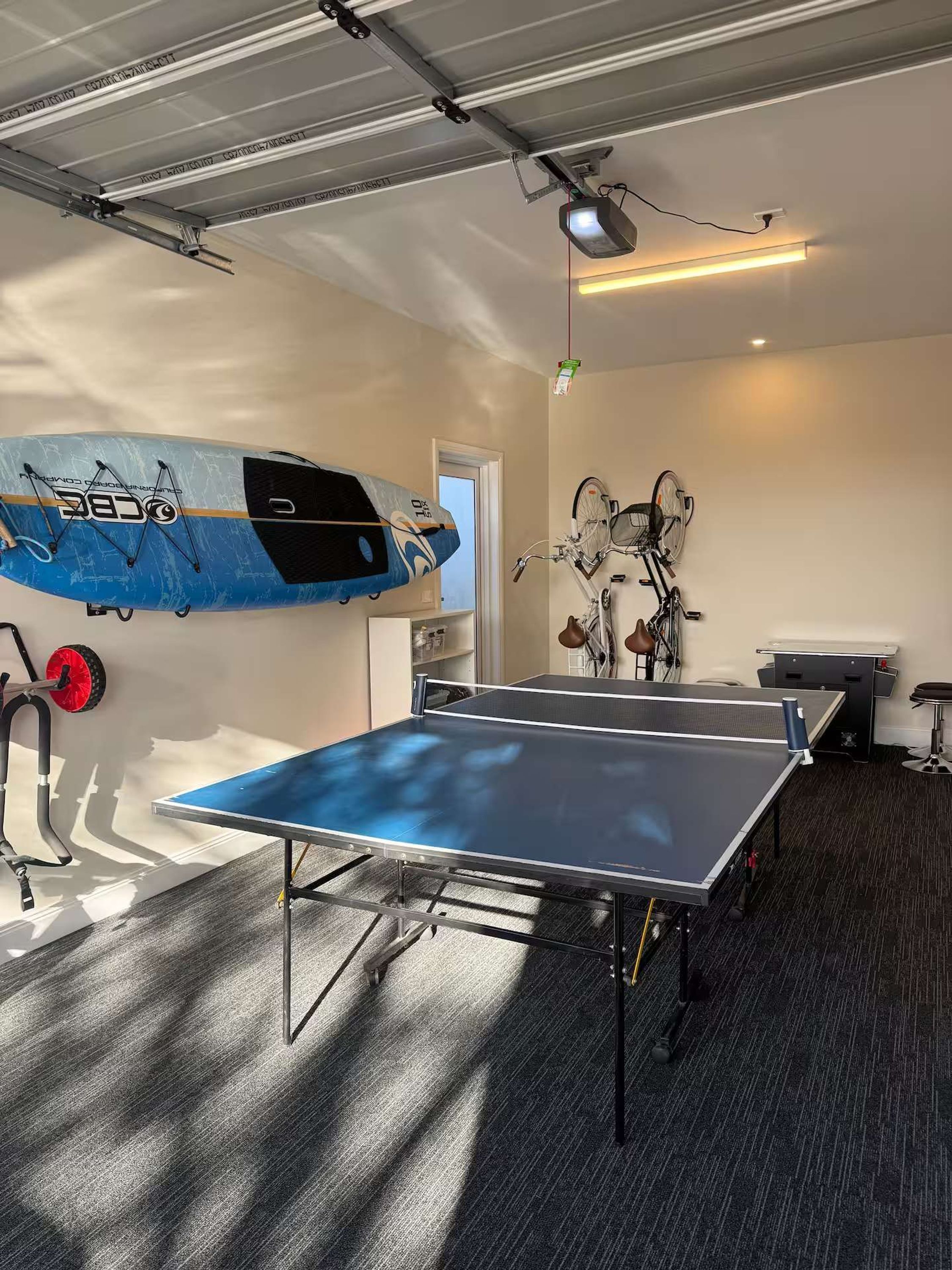 A room with a ping pong table, a bicycle, and a surfboard.