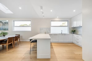 A modern kitchen with white cabinetry, a central island, and a dining area with wooden chairs and a table. - Oakwood Retreat in Rosebud (Melbourne)