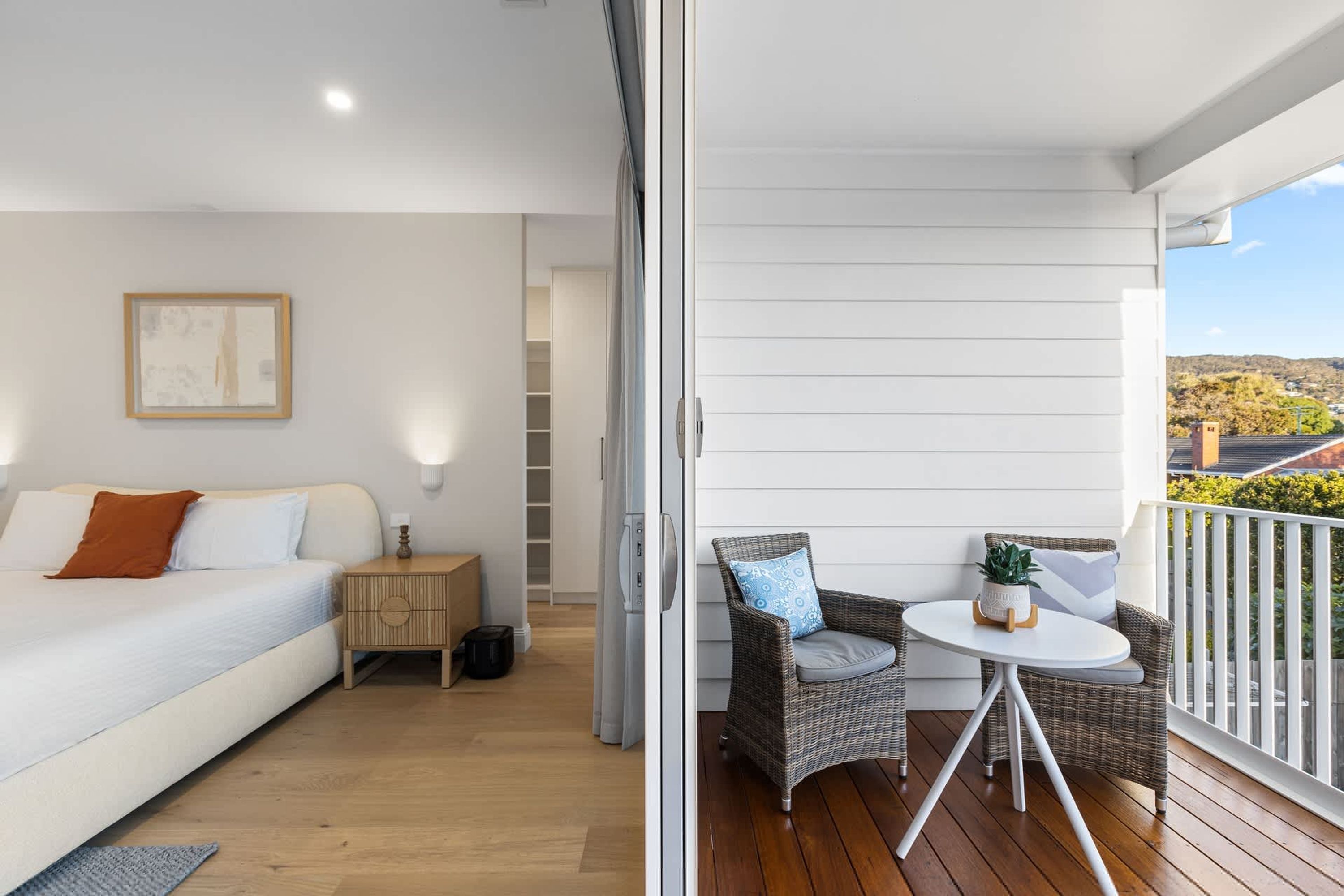 A bedroom with a bed, bedside table, and a nightstand. There is a balcony with wicker furniture and a potted plant.