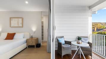 A bedroom with a bed, bedside table, and a nightstand. There is a balcony with wicker furniture and a potted plant.