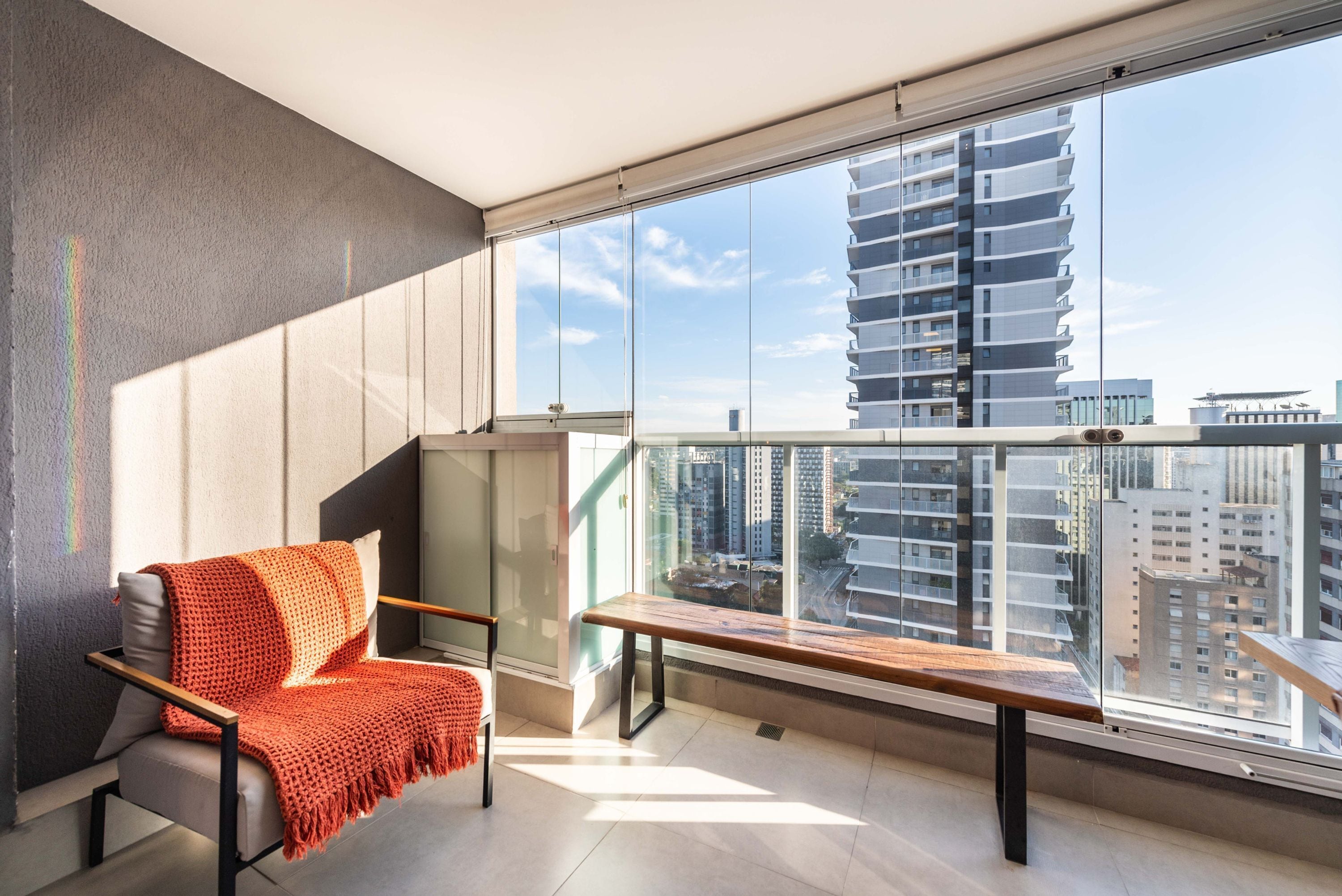 Een modern balkon met uitzicht op hoge gebouwen, een grijze bank met een rode deken en een houten bankje.