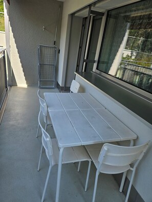 A balcony with a white table and chairs, a metal railing, and a view of a building through a window. - Parkareal Davos Vacation Rental for 2 People: Comfortable, Cozy, and Centrally Located (Davos Platz)