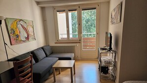 A living room with a sofa, a wooden table, a chair, a radiator, a television on a stand, and a window with a view of greenery. - Parkareal Davos Vacation Rental for 2 People: Comfortable, Cozy, and Centrally Located (Davos Platz)