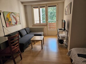 A living room with a sofa, a wooden table, a chair, a radiator, a television on a stand, and a window with a view of greenery. - Parkareal Davos Vacation Rental for 2 People: Comfortable, Cozy, and Centrally Located (Davos Platz)