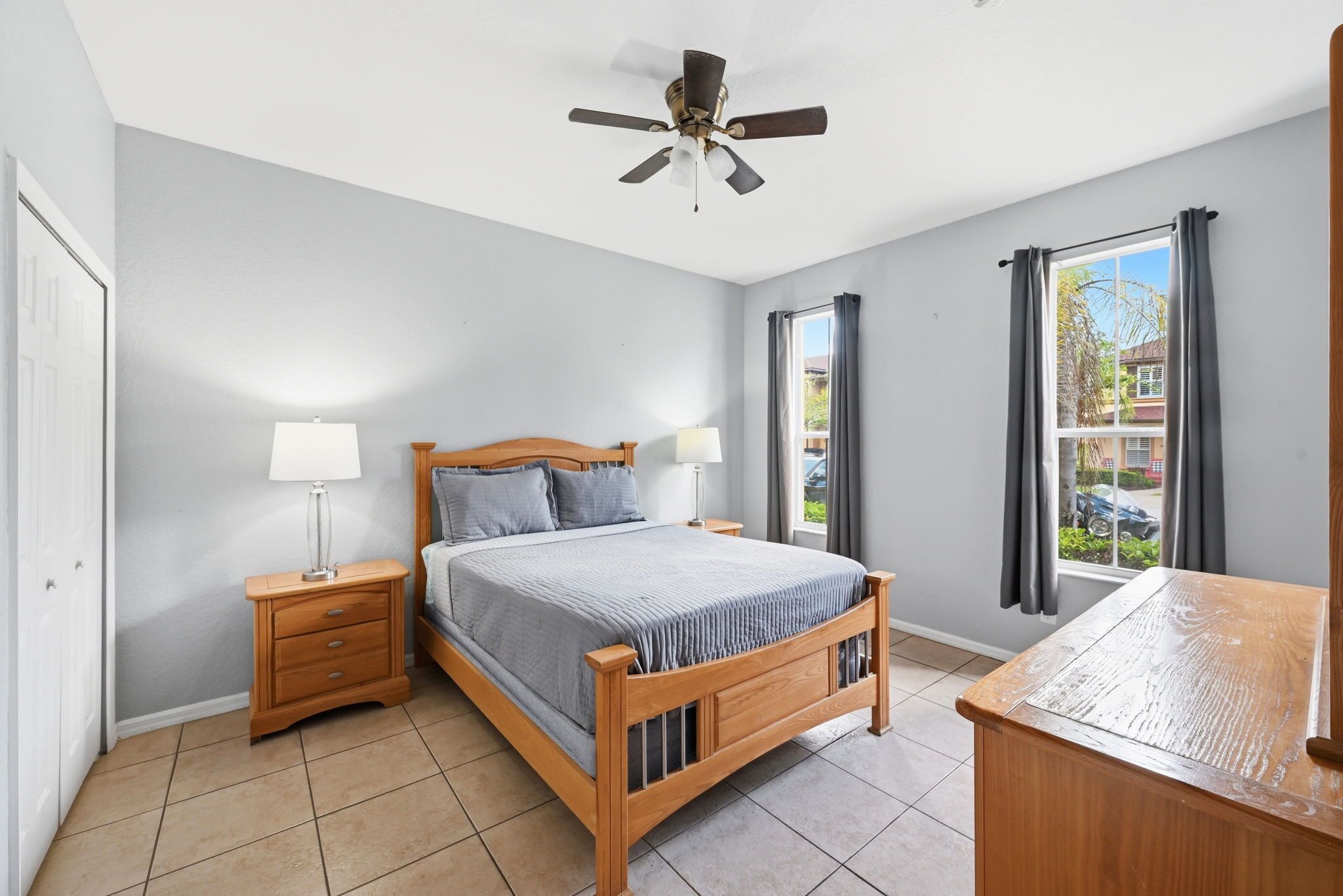 Un dormitorio con cama, mesitas de noche, ventilador de techo, ventana con cortinas y vistas al exterior.