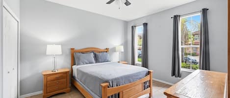 Un dormitorio con cama, mesitas de noche, ventilador de techo, ventana con cortinas y vistas al exterior.
