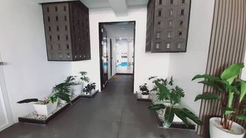 A hallway with potted plants, a view into another room, and a large window with blinds.