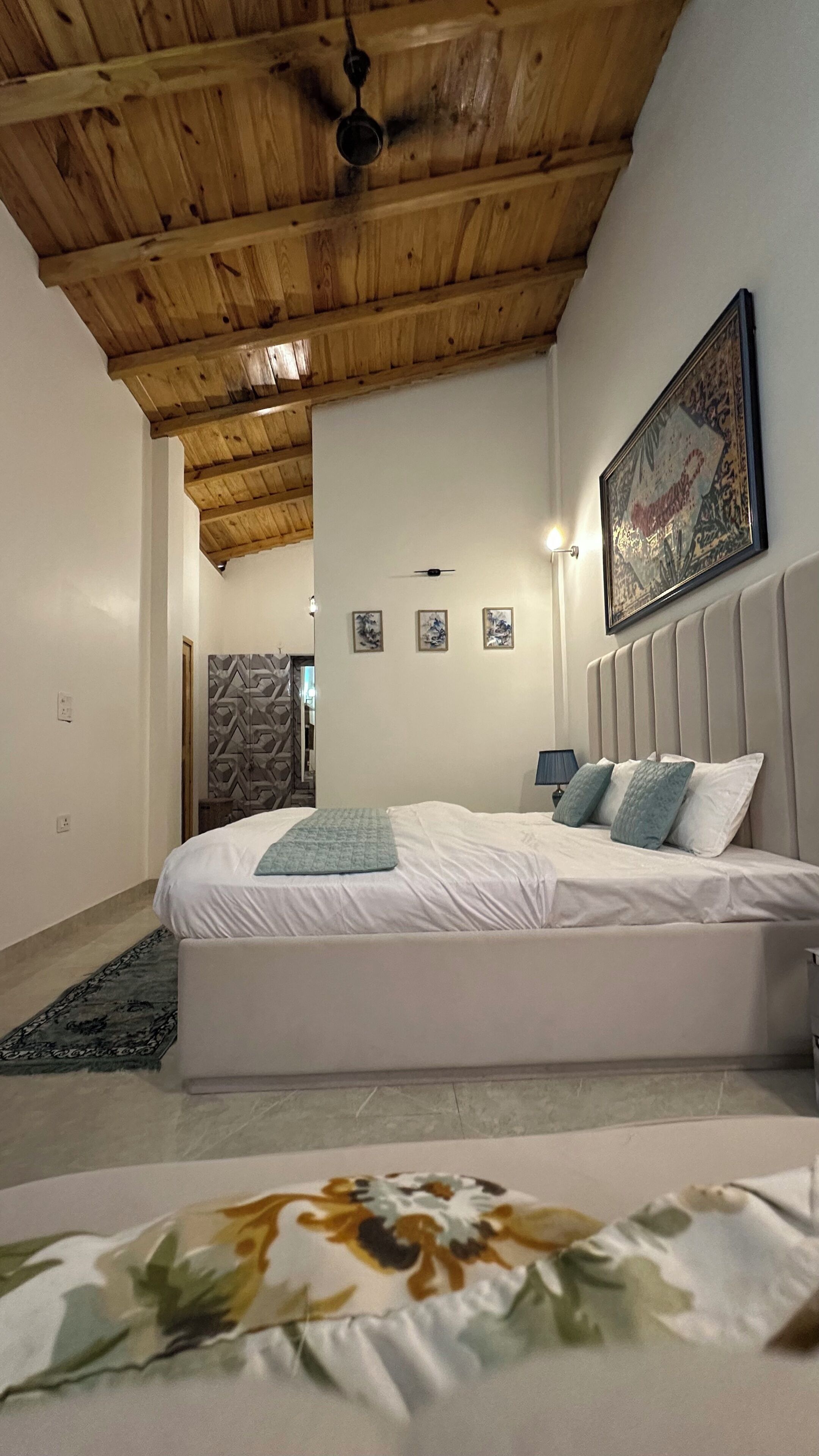 A bedroom with a wooden ceiling, a bed with white bedding, a headboard, a wall-mounted lamp, and a framed picture.