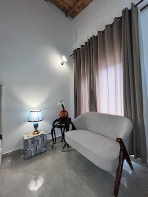 Una habitación con sofá, lámpara, una mesita con un jarrón y ventana con cortinas.