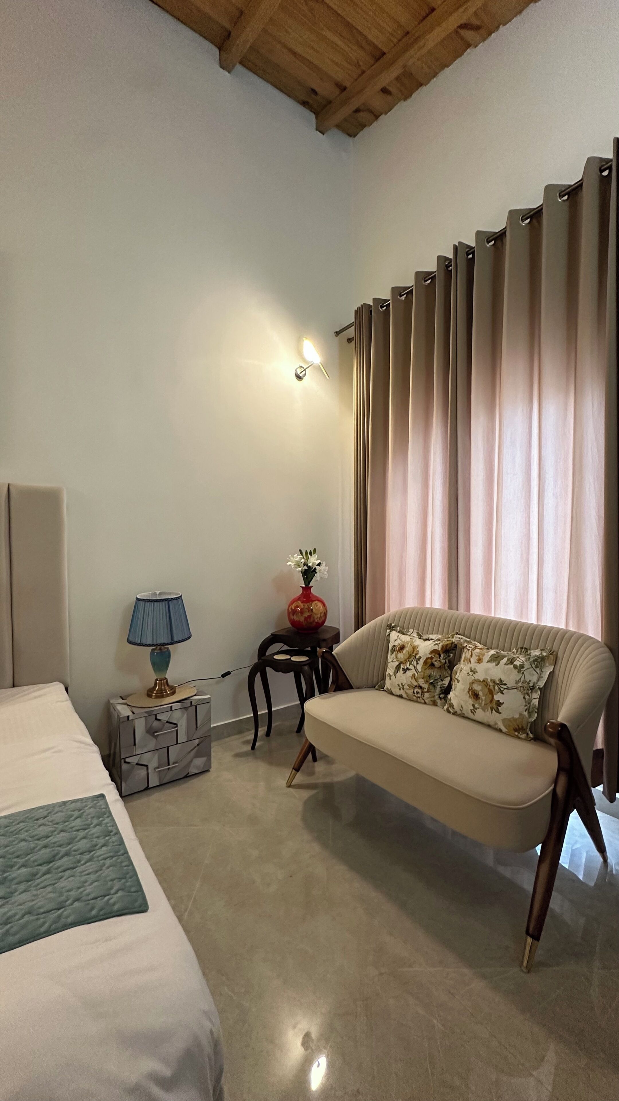 A hotel room with a bed, a sofa, a lamp, a vase with flowers, and a curtain.