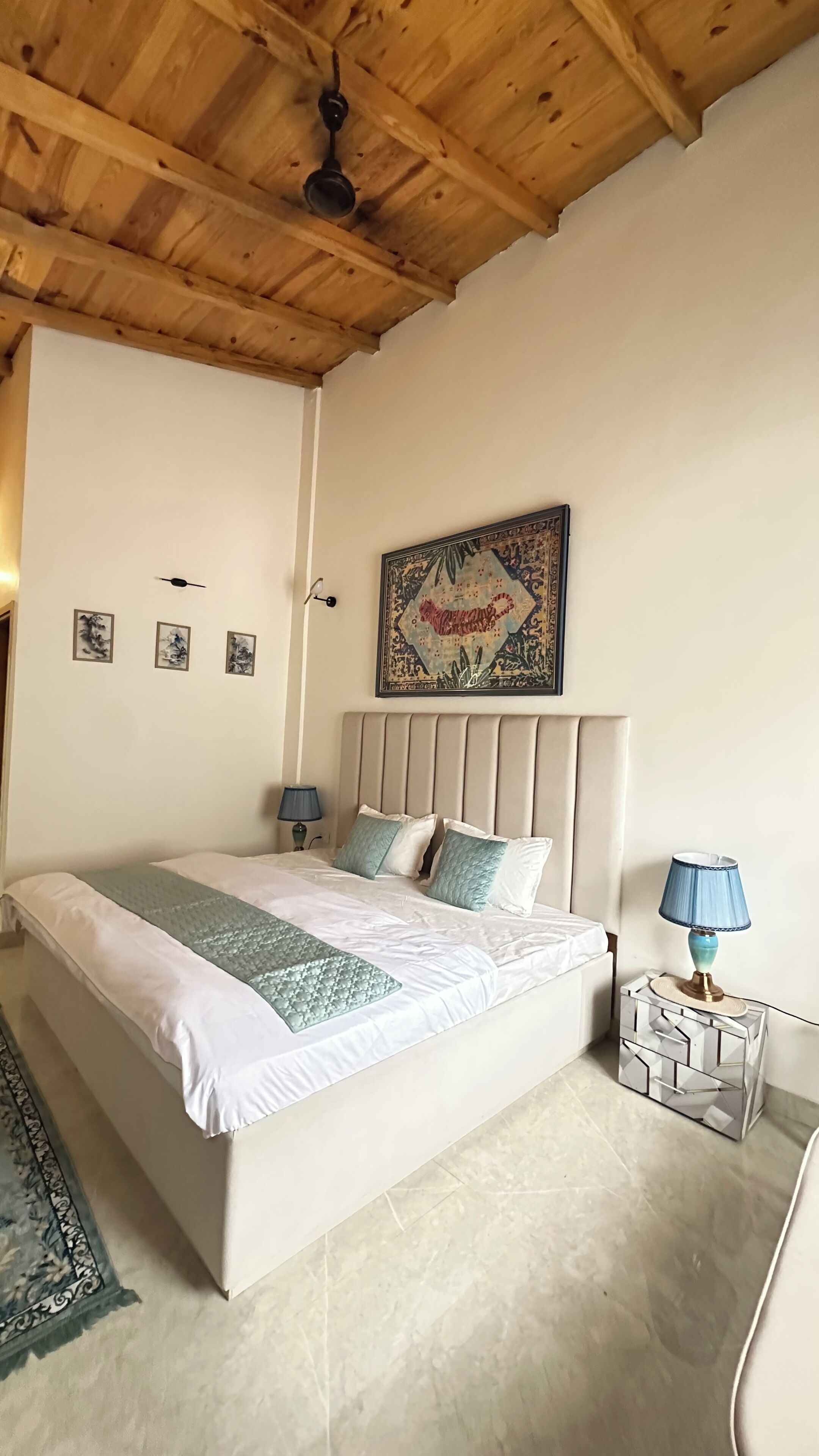 A bedroom with a wooden ceiling, a bed with white bedding and blue pillows, a decorative rug, a wall-mounted lamp, and a framed artwork above the bed.