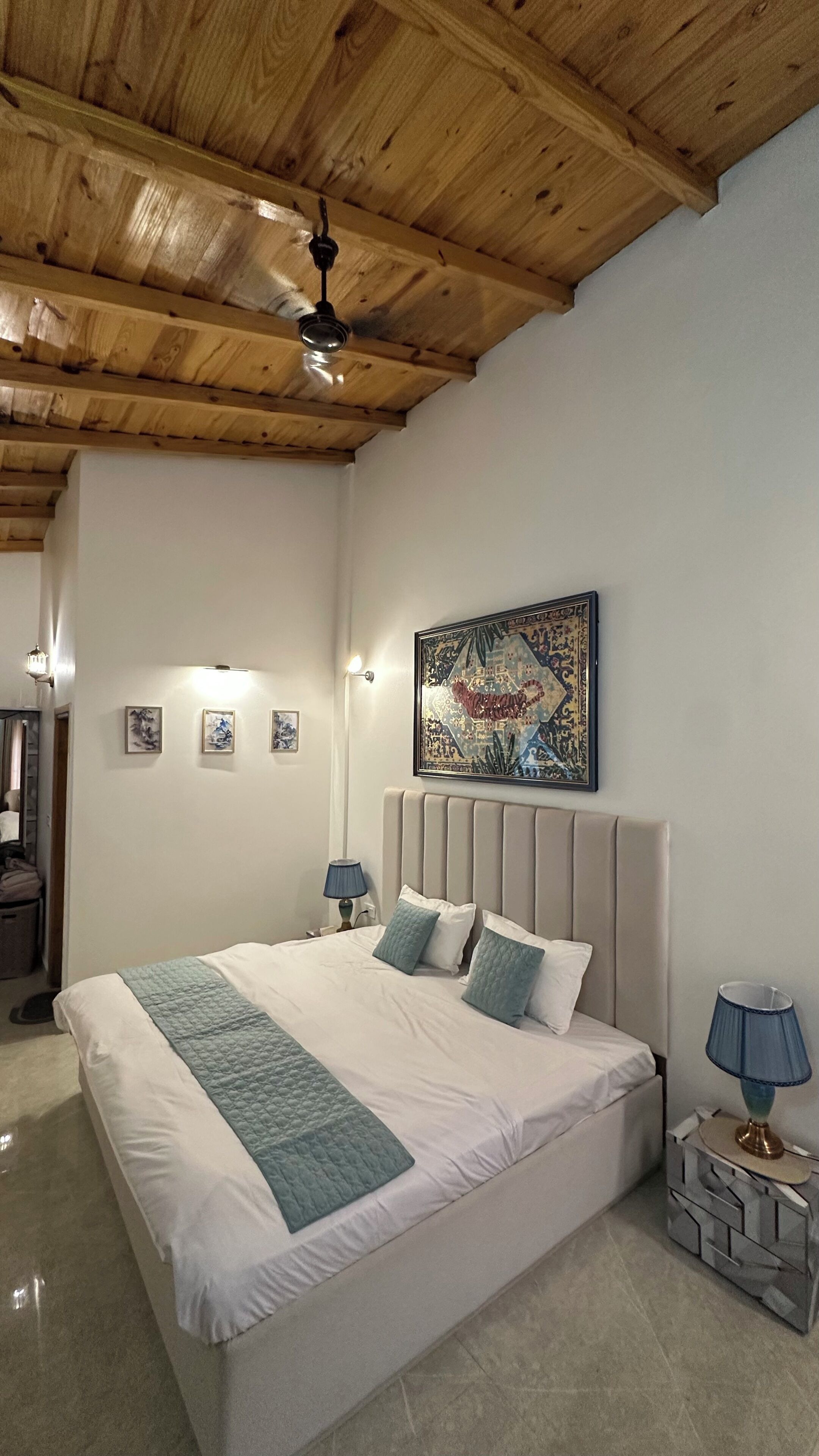 A bedroom with a wooden ceiling, a bed with white bedding and blue pillows, a bedside lamp, and a wall-mounted painting.