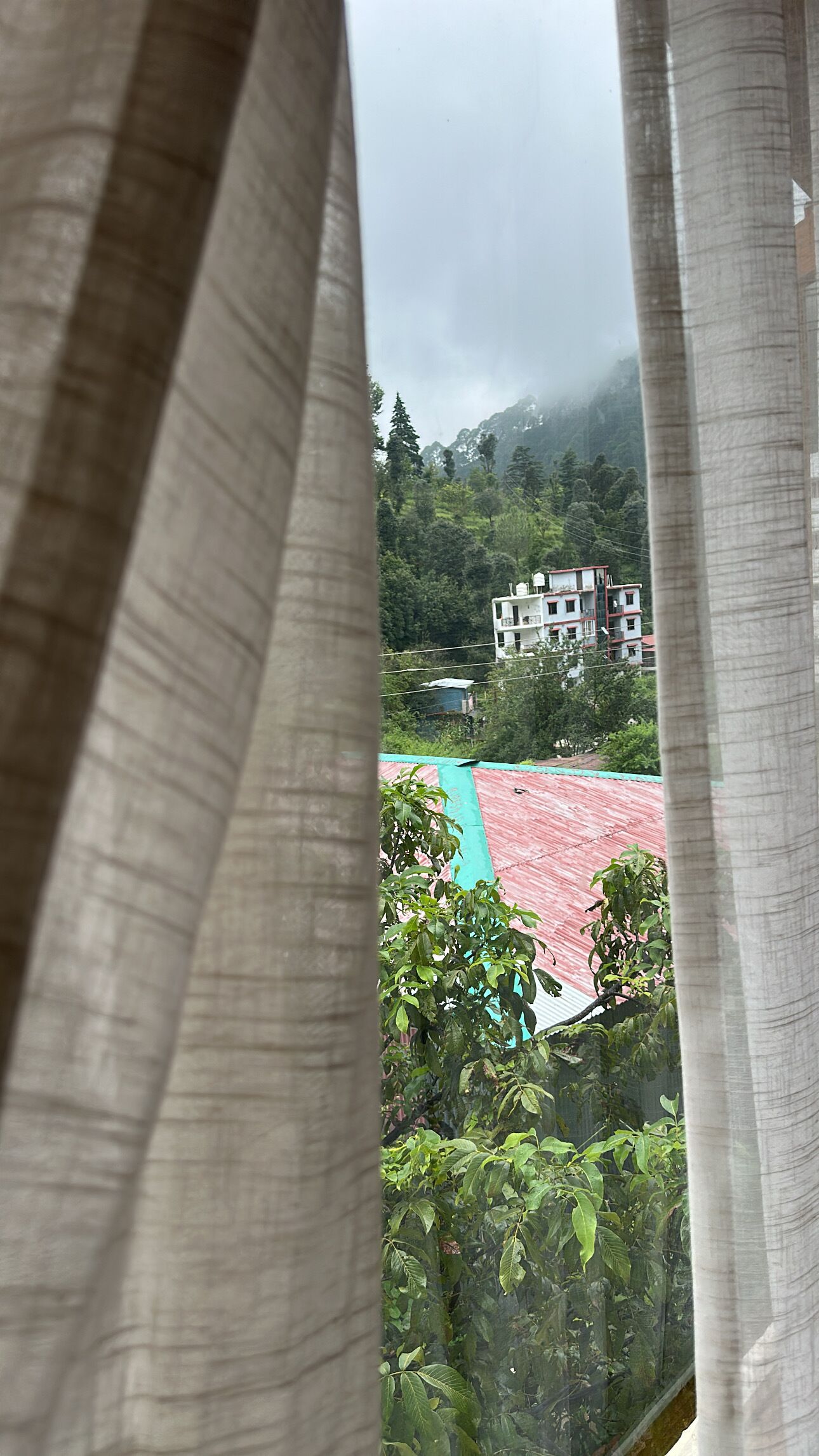 Une vue à travers une fenêtre avec des rideaux, montrant un bâtiment avec un toit rouge et un champ verdoyant au premier plan.