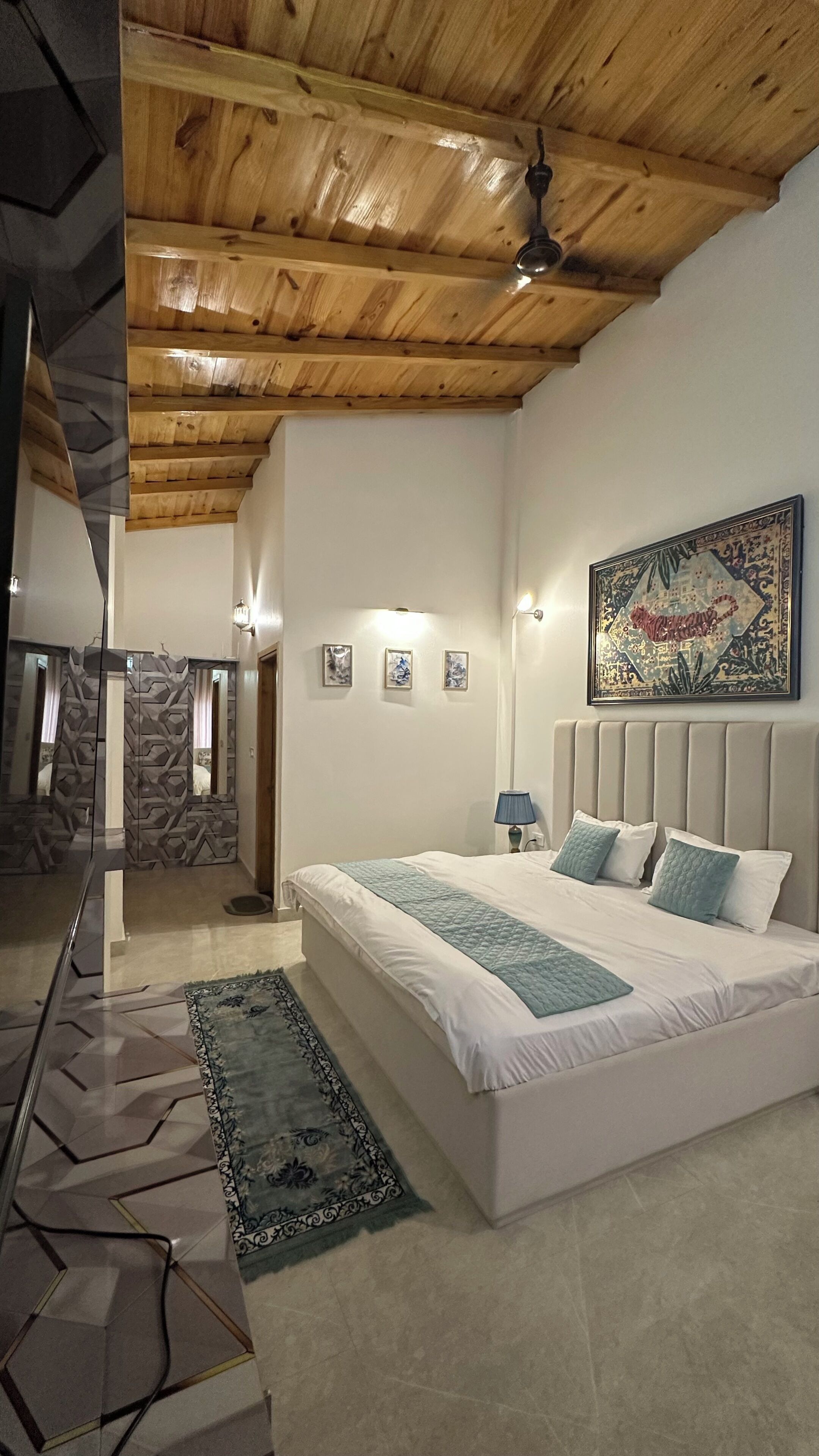 A bedroom with a large bed, a ceiling fan, a wall-mounted lamp, and a painting.