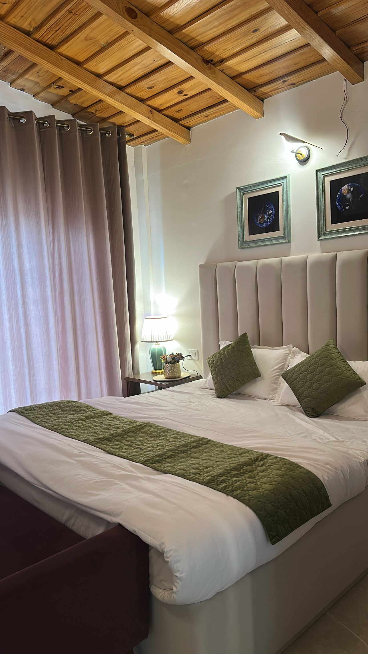 Una camera d'albergo con soffitto in legno, un letto con biancheria verde e bianca, un comodino con una lampada e due quadri appesi al muro.