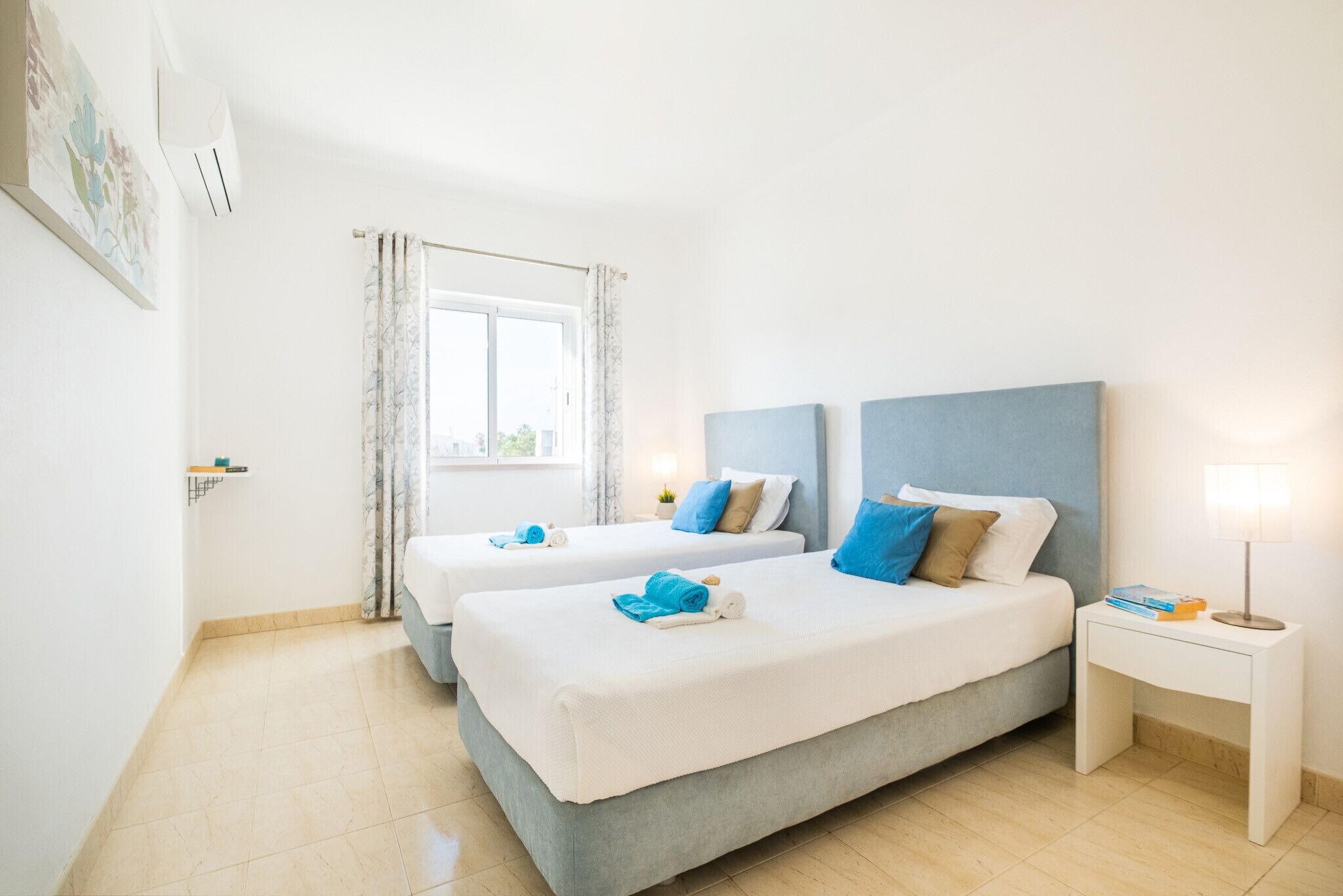 A hotel room with two beds, a window with curtains, a bedside table with a lamp, and a wall-mounted air conditioner.