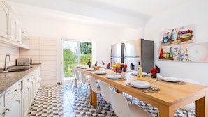 A modern kitchen with a dining area, featuring a long wooden table set for six, white chairs, and a view of greenery through the glass door. - Holiday villa with BBQ and heatable pool (Albufeira)