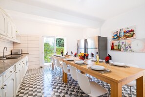 A modern kitchen with a dining area, featuring a long wooden table set for six, white chairs, and a view of greenery through the glass door. - Holiday villa with BBQ and heatable pool (Albufeira)