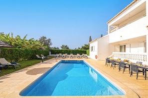 A swimming pool with lounge chairs and tables, surrounded by a white building with a tiled roof and a hedge. - Holiday villa with BBQ and heatable pool (Albufeira)
