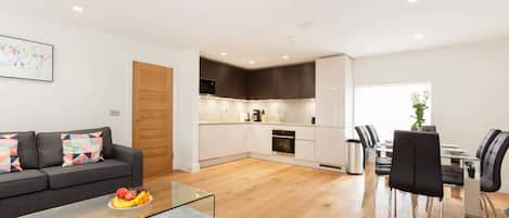 A modern living space with a sofa, a glass coffee table with fruit, a kitchen area, and a dining table with chairs.