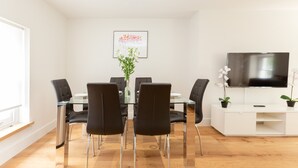 Ein moderner Essbereich mit Glastisch, schwarzen Stühlen, einer Blumenvase, einem gerahmten Bild an der Wand und einem Fernseher auf einem weißen Schrank.