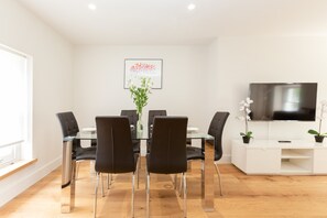 Ein moderner Essbereich mit Glastisch, schwarzen Stühlen, einer Blumenvase, einem gerahmten Bild an der Wand und einem Fernseher auf einem weißen Schrank.