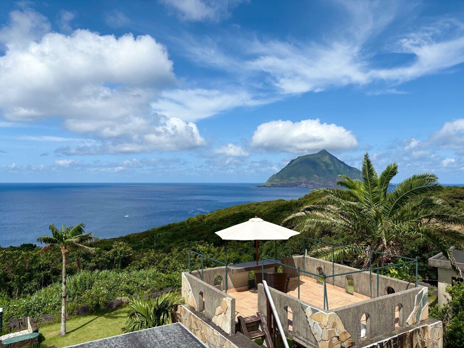 Um terraço com mesa e cadeiras, com vista para uma paisagem verdejante e o oceano.