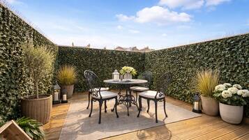 A patio with a round table and chairs, surrounded by potted plants and a decorative hedge.