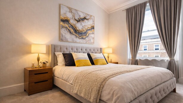 Une chambre à coucher avec un grand lit, deux tables de chevet, une tête de lit, une fenêtre avec des rideaux et une œuvre d’art accrochée au mur.