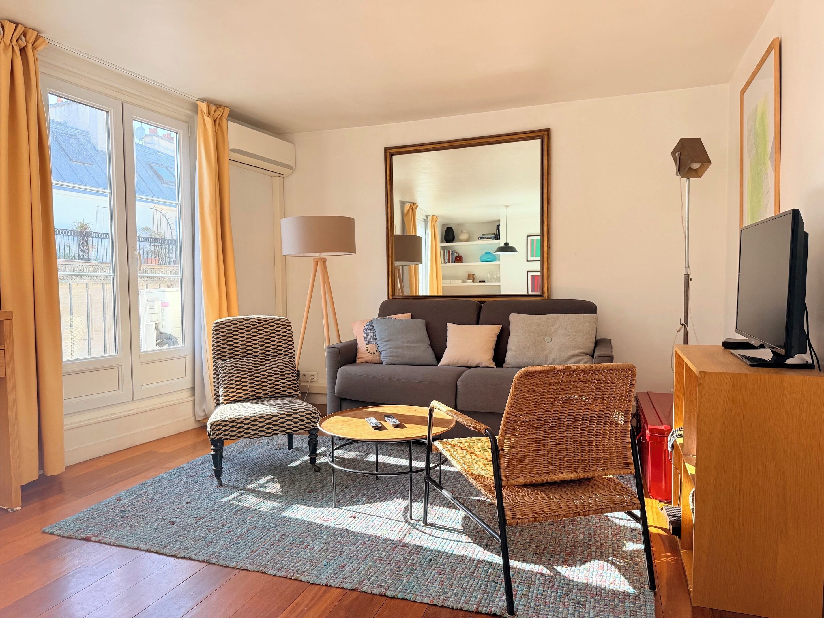 A living room with a sofa, two armchairs, a coffee table, a TV on a wooden stand, a floor lamp, and a large mirror on the wall.
