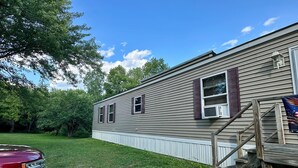 A single-story mobile home with a front porch, a small staircase, and a vehicle parked in the yard.