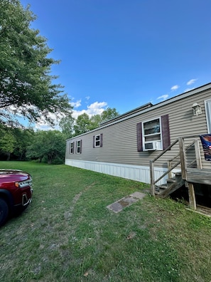 A single-story mobile home with a front porch, a small staircase, and a vehicle parked in the yard.