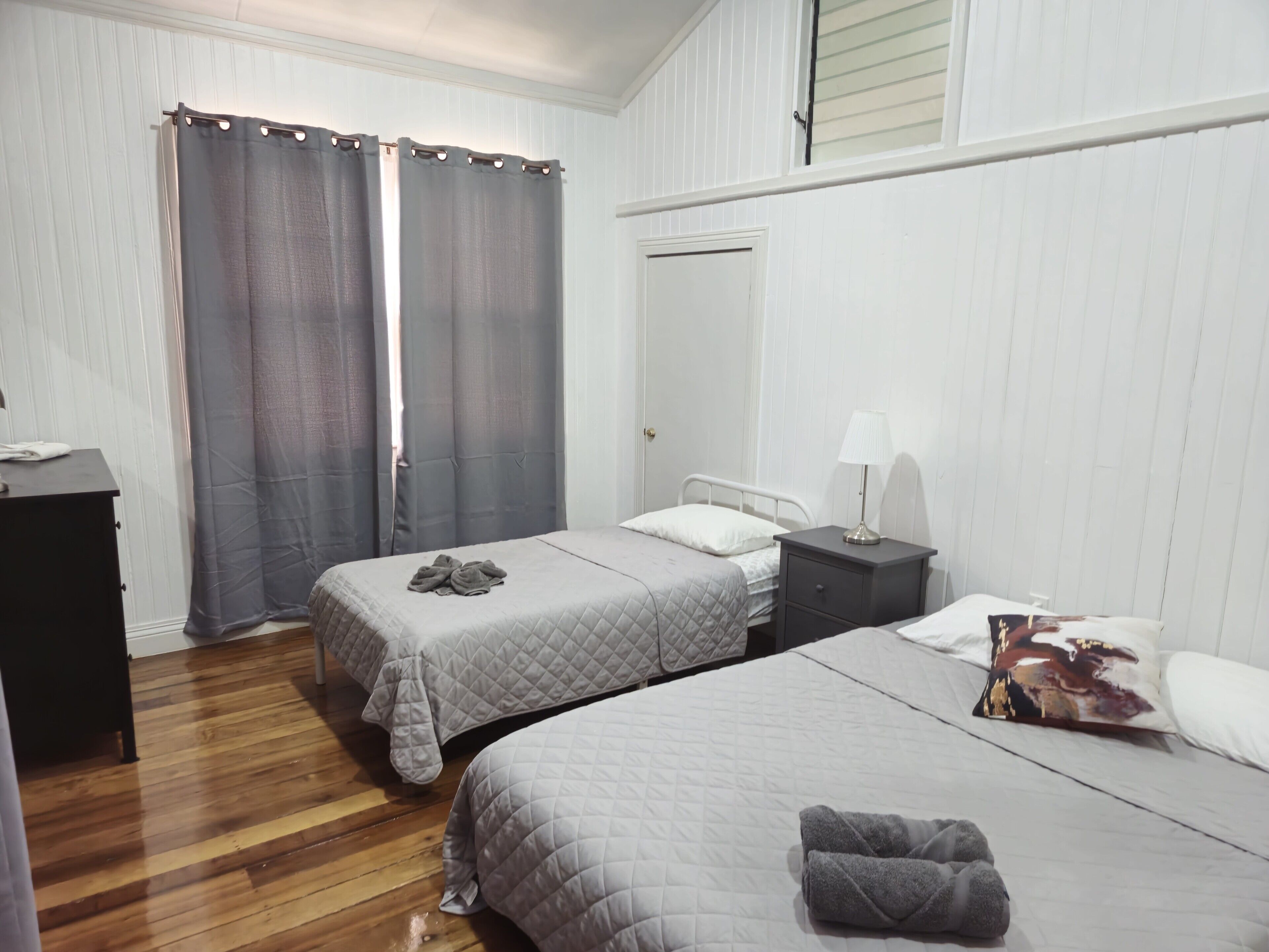 A hotel room with two beds, a nightstand with a lamp, a wardrobe, and a window with curtains.