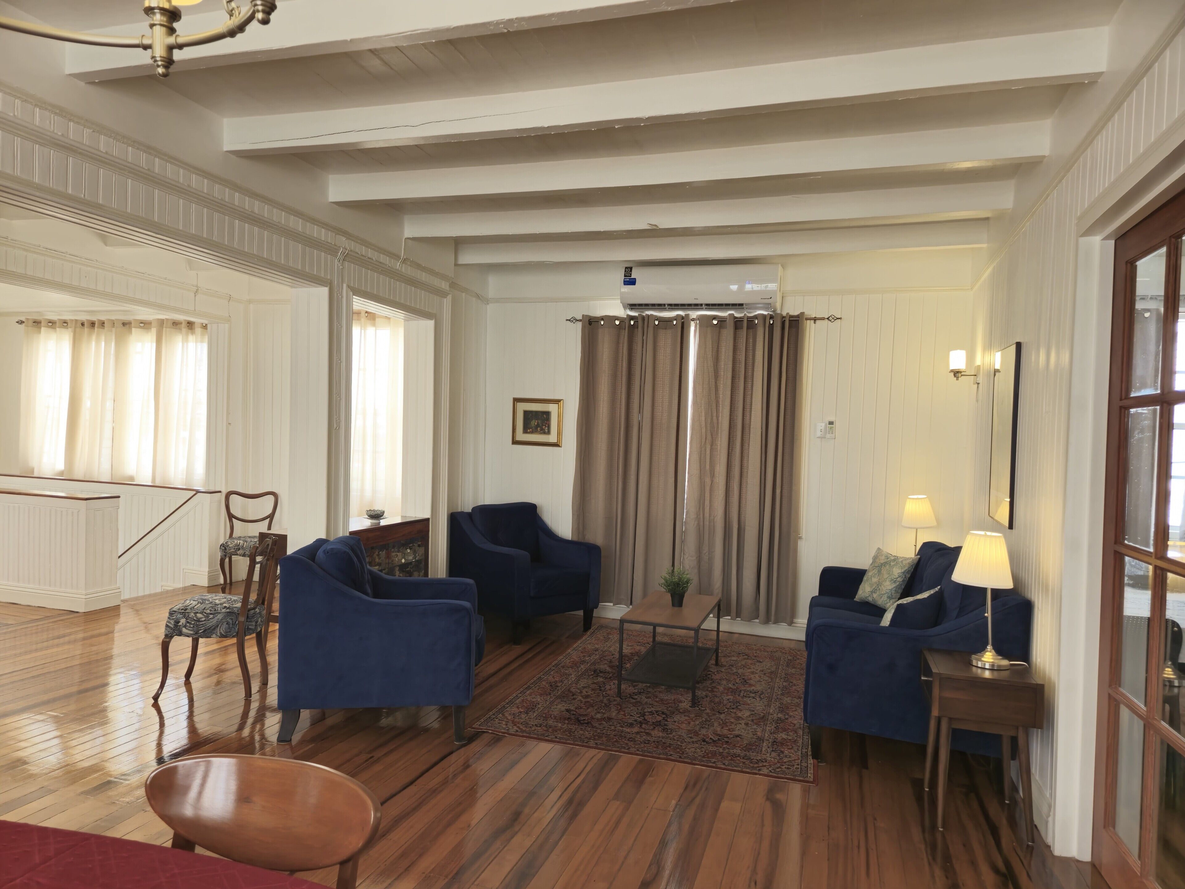 A spacious living room with a high ceiling, a wooden floor, and a large window with curtains. There are two blue armchairs, a wooden coffee table, and a small side table with a lamp.
