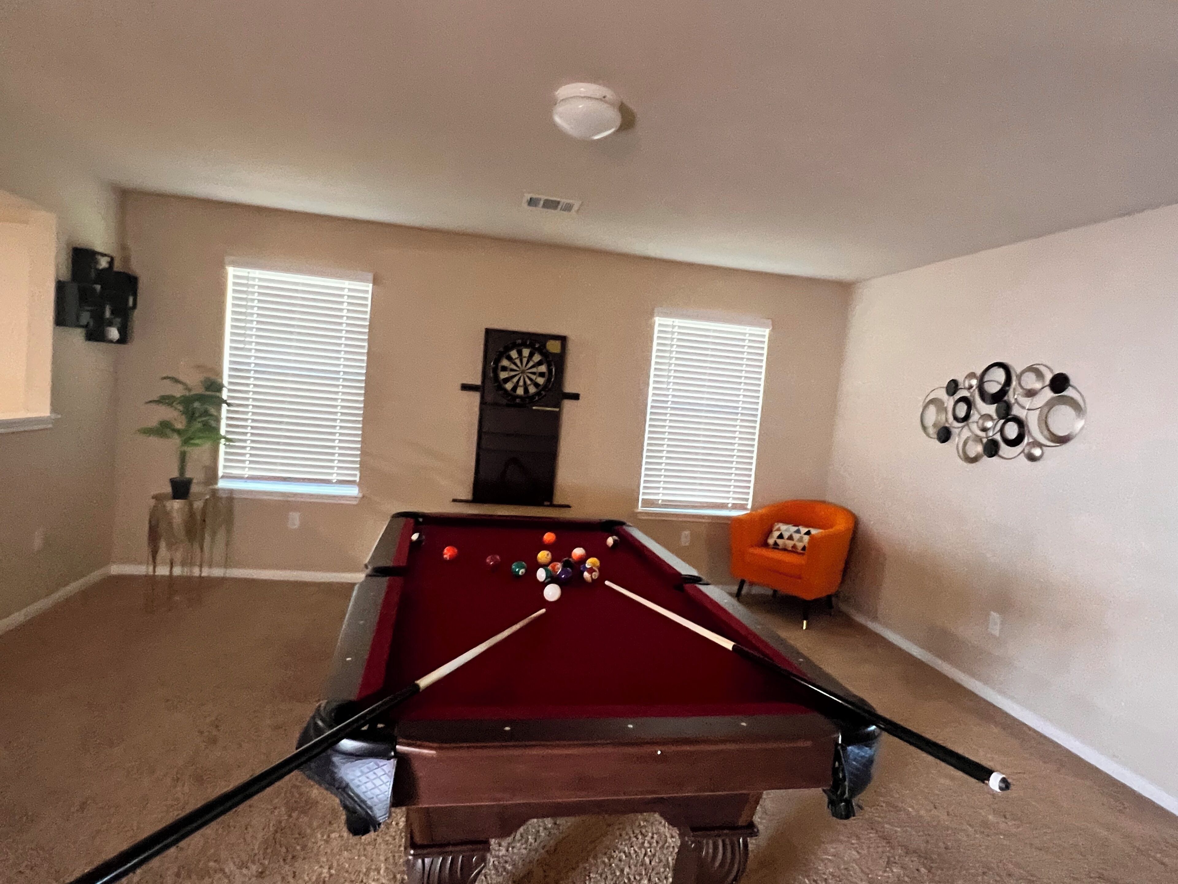 Une table de billard dans une pièce dotée de deux fenêtres, d’un jeu de fléchettes et d’un élément décoratif fixé au mur.
