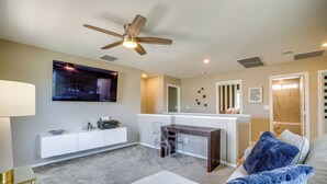 A modern living room with a ceiling fan, a flat-screen TV mounted on the wall, a desk with a chair, and a cozy seating area with a blue blanket. - Stylish Family Home w/ Playground in Las Vegas (Las Vegas)