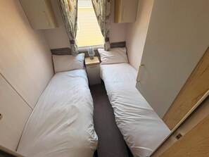 Ein schmales Zimmer mit zwei Betten, einem kleinen Tisch und einem Fenster mit Vorhängen.