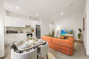A modern kitchen with white cabinets, a dining area with a table set for four, and an orange sofa in a living area with a TV displaying a beach scene. - Walk to Central |Boutique Retreat in Surry Hills (Surry Hills)