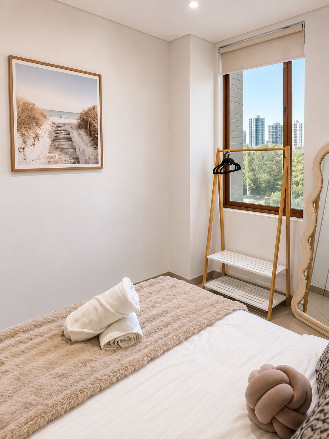A bedroom with a bed, a fluffy rug, a towel, a picture on the wall, a window with a view of buildings, and a wooden hanger stand.