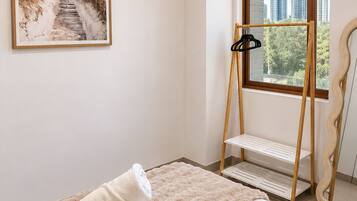 A bedroom with a bed, a fluffy rug, a towel, a picture on the wall, a window with a view of buildings, and a wooden hanger stand.