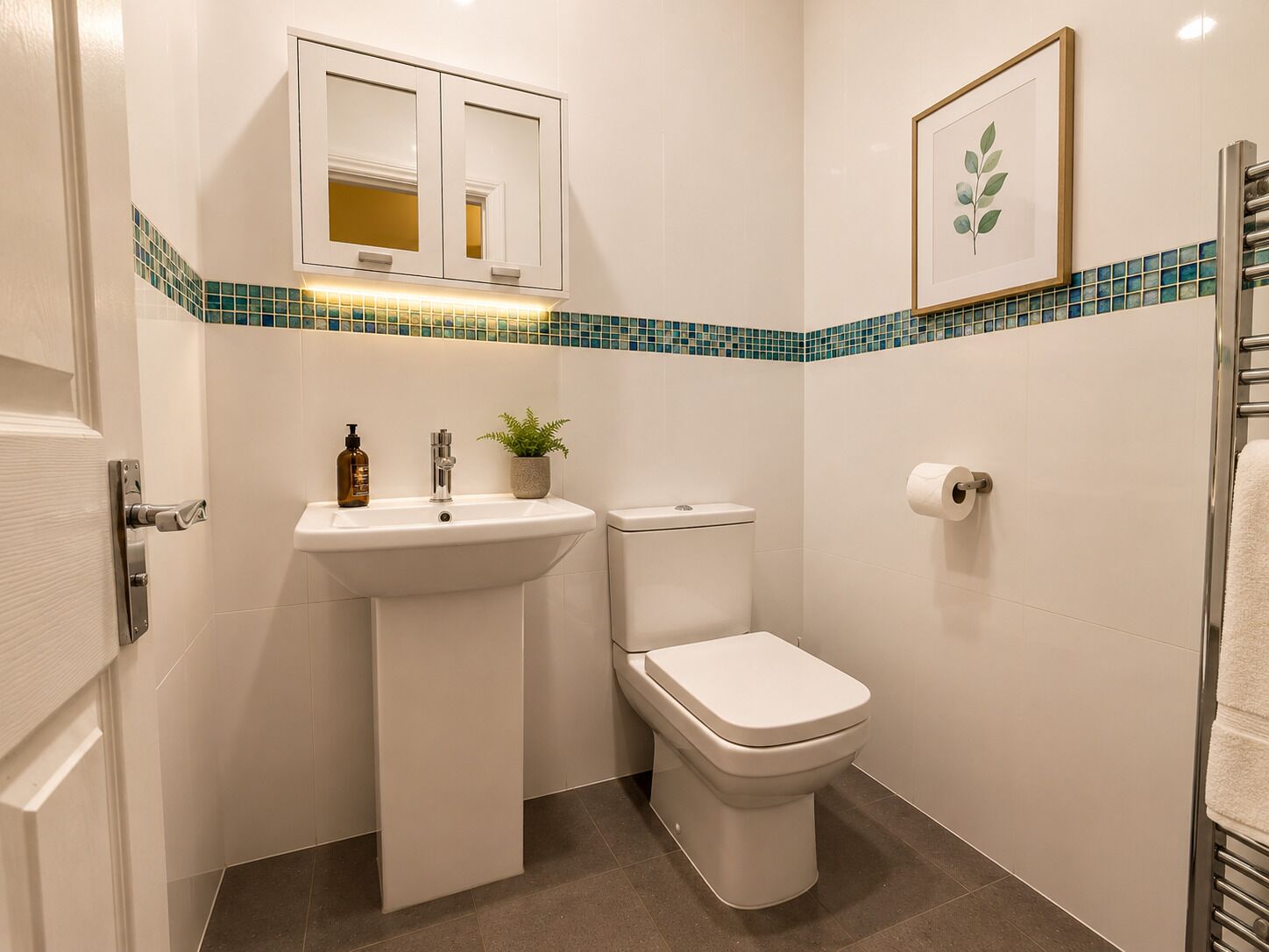 Um banheiro compacto com pia branca, vaso sanitário, pequeno espelho sobre a pia, quadro na parede e porta-toalhas.
