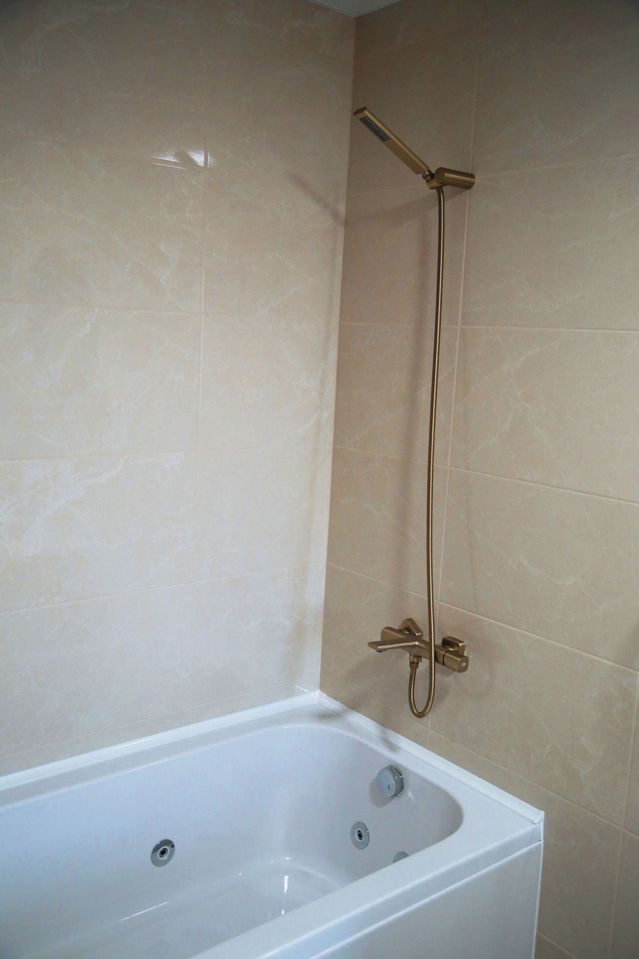 Une salle de bain avec une baignoire blanche, une douche dotée d’un pommeau de douche doré et des murs recouverts de carreaux beiges.