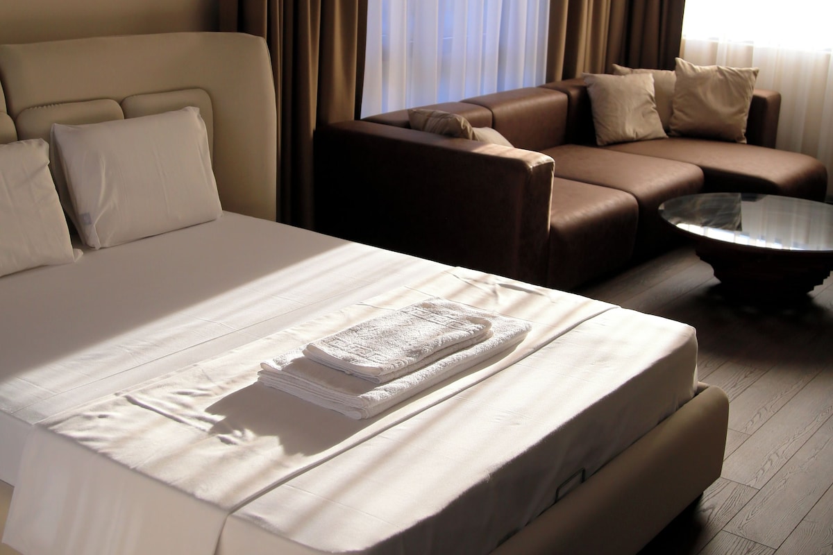 Habitación de hotel con una cama grande, un sofá, una mesita y una ventana con cortinas.