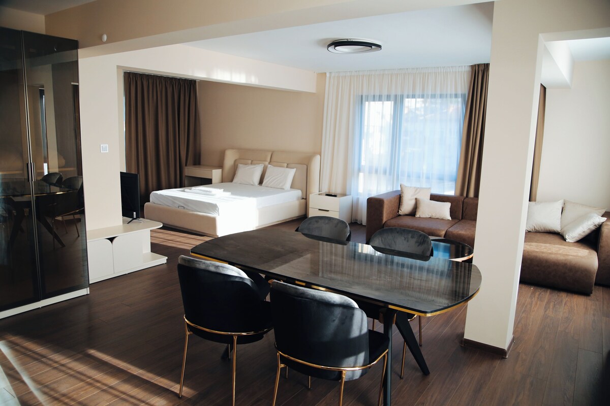 A modern hotel room with a bed, a sofa, a dining table with chairs, and a television.