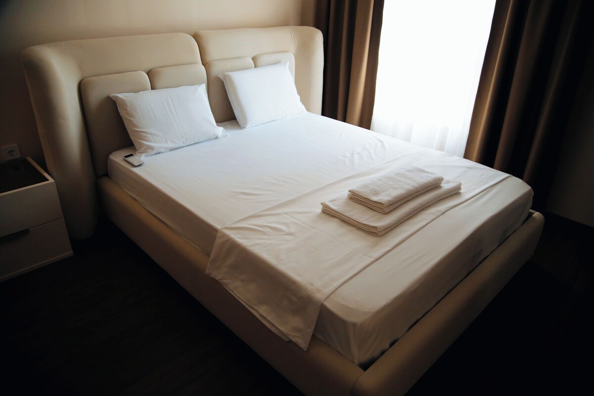 A neatly made bed with white linens and pillows, a bedside table with a lamp, and a window with curtains.