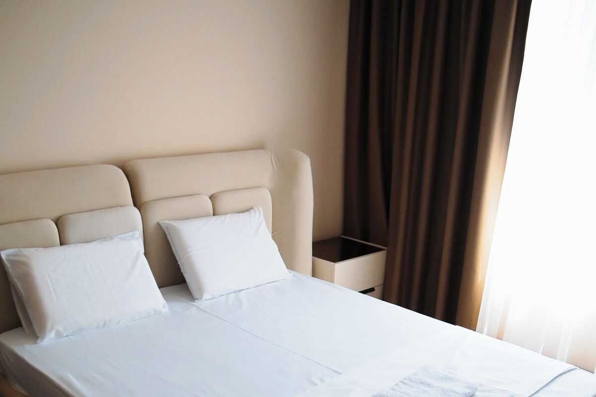 Una cama bien hecha con sábanas y almohadas blancas, una mesita de noche con lámpara y una ventana con cortinas.