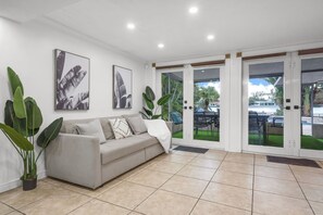 A living room with a sofa, two large potted plants, and a view through glass doors to an outdoor area with a pool and chairs. - Waterfront Jungle Home with Kayaks (Miami)